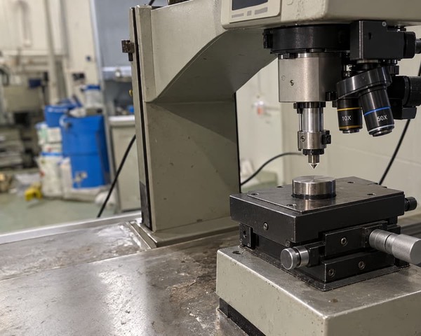 Benchtop hardness tester with its indenter head lowered toward a steel test sample on the anvil