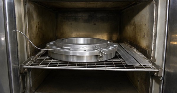 Heavy nickel plated steel flange with a thermocouple clamped to its rim beside two thin plated test strips on an oven shelf